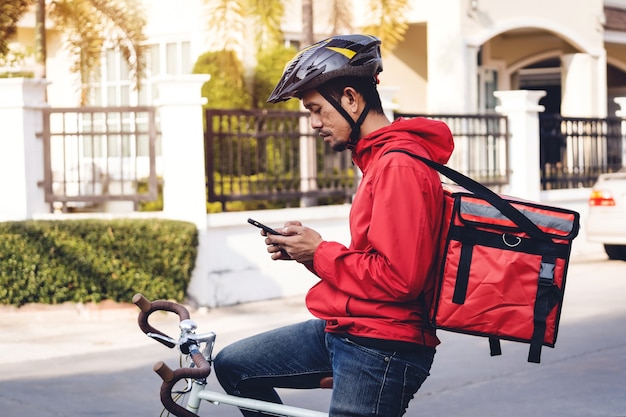 Koerier in rood uniform met een bezorgdoos op de fiets en op de mobiele telefoon kijkend om het adres te controleren om eten bij de klant te bezorgen. Koerier op een fiets die voedsel in de stad bezorgt.