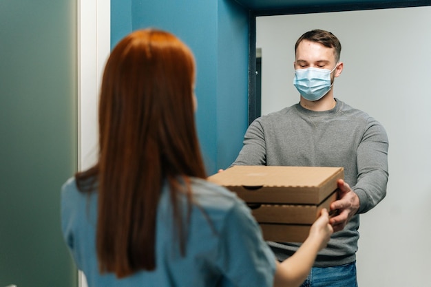 Koeriersman met medisch gezichtsmasker die dozen met hete pizza bezorgt aan onherkenbare vrouwelijke klant voor de deur thuis