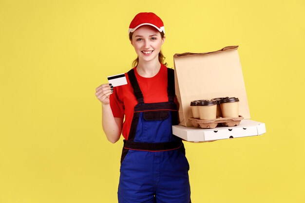 Koeriersvrouw met creditcard en koffie met pizzadoos die naar de camera kijkt met een vrolijke uitdrukking