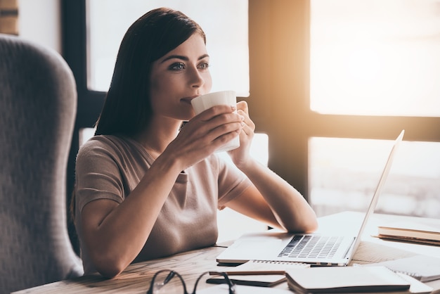 Koffie brengt haar inspiratie. Jonge mooie vrouw die koffie drinkt en wegkijkt terwijl ze in een stoel op haar werkplek zit