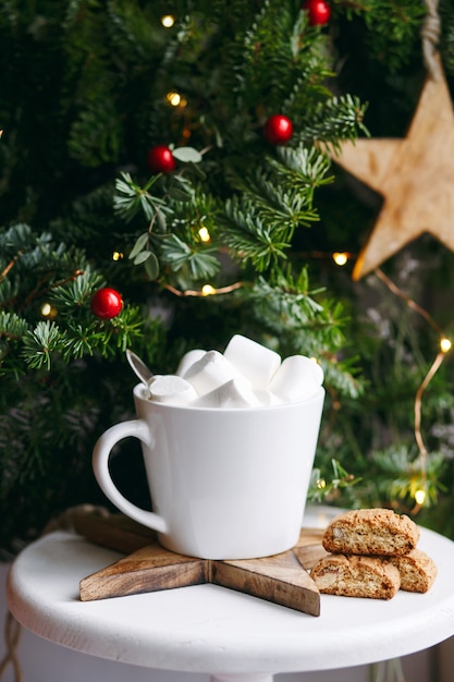 Koffie in een witte kop met marshmallows. Feestelijke ochtendkoffie met traditionele Italiaanse cantuccini-amandelkoekjes. Een kopje koffie op een achtergrond van groene dennentakken op een witte standaard.