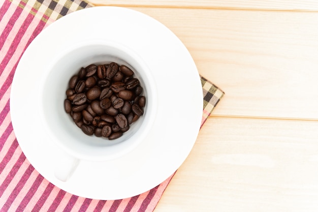 Koffiebonen in witte kop, stoffenplaid op een houten tafel.