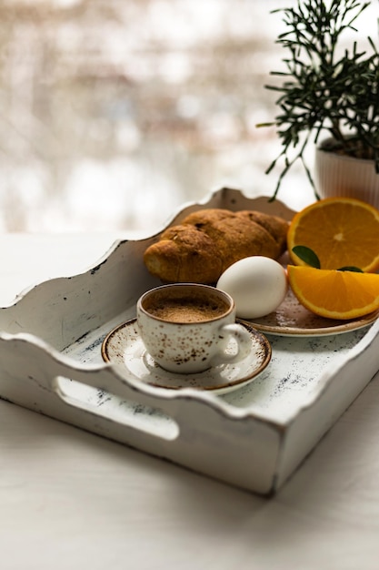 Koffiekopje croissant ei vers oranje fruit op wit houten dienblad tegen wazig venster achtergrond