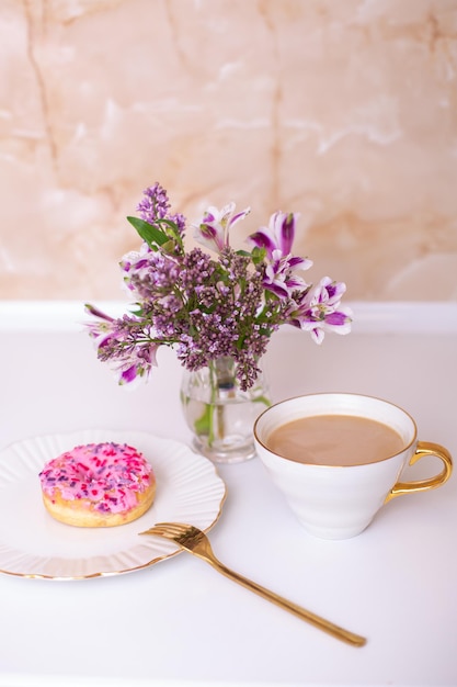 Koffiekopje en zoete aardbeidoughnut op gouden plaat met bloemboeket