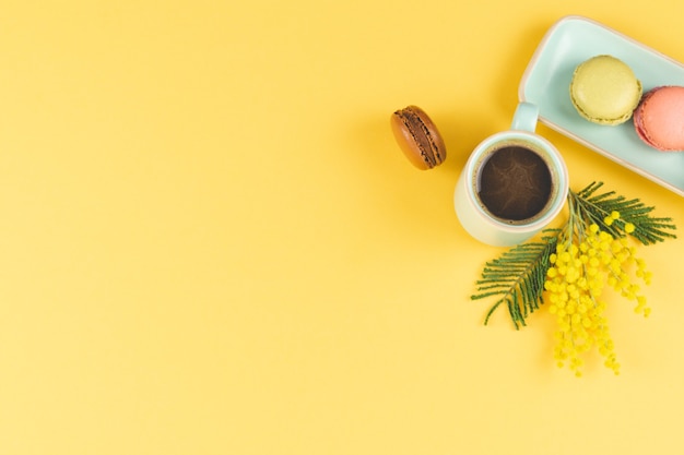 Koffiemok met macarons en gele bloemendecoratie op geel. Bovenaanzicht.