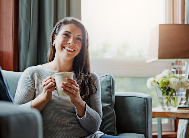 Koffievrouw en glimlach van een ontspannen persoon thuis op een bank in de woonkamer ontspannend in de ochtend Denkende gelukkige vrouw en lensflare op een bank in een loungehuis met thee geluk voelen met een mok