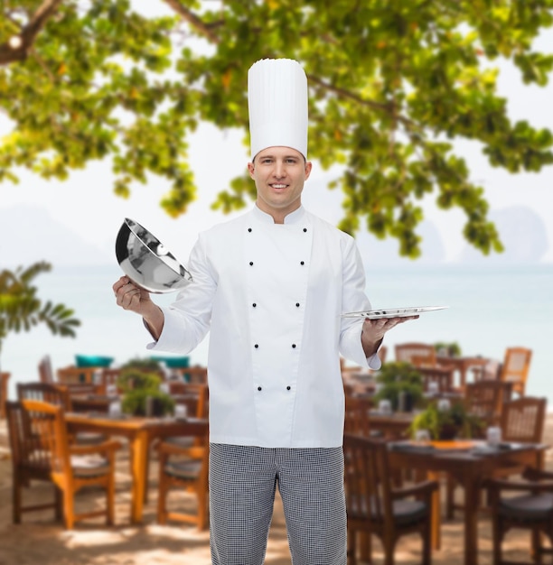koken, beroep en mensenconcept - gelukkige mannelijke chef-kok die clochedekking opent over restaurantlounge op strand