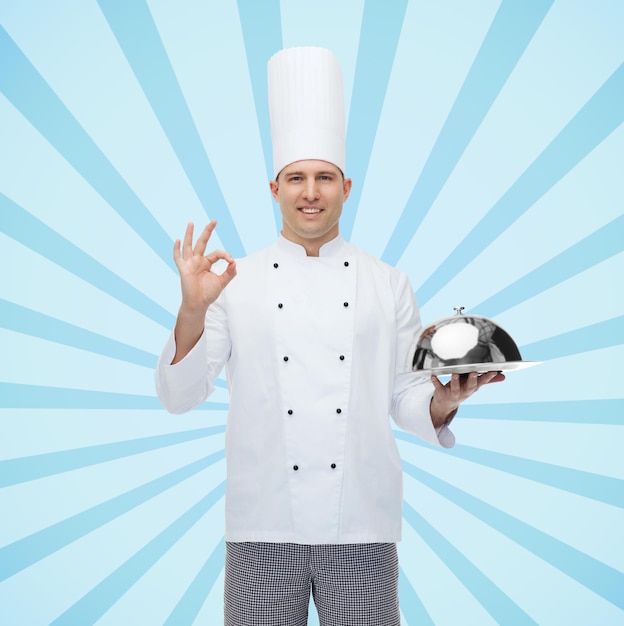 koken, beroep, gebaar en mensenconcept - gelukkige mannelijke chef-kok die cloche houdt en ok teken toont over blauwe burst-stralenachtergrond