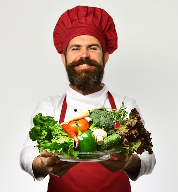 Koken en vegetarisch dieet concept Chef houdt kom met verse groenten Man met baard geïsoleerd op witte achtergrond Kok met trotse glimlach in bordeaux uniform houdt salade ingrediënten