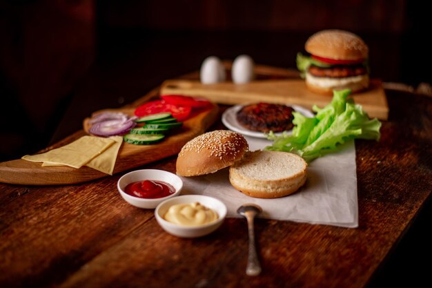 Koken hamburger of cheeseburger Verschillende ingrediënten voor een klassieke hamburger Gegrild vlees groenten Groenen sauzen in de buurt van een sesambroodje Houten achtergrond Hamburger dag