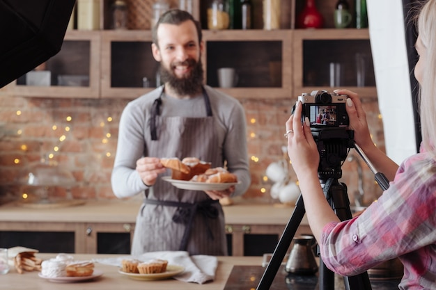 Koken kerel. Bakken. Opgewonden chef-kok in schort met plaat van verse taarten en gebak. Backstage fotografie.