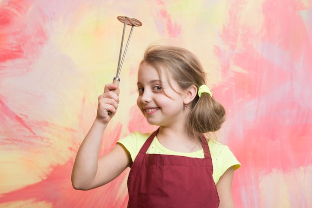 Koken met keukengereedschap. Jeugd en opvoeding. Kid in kok schort. Klein meisje op kleurrijke achtergrond. Chef-kokkind met stamper voor aardappel.
