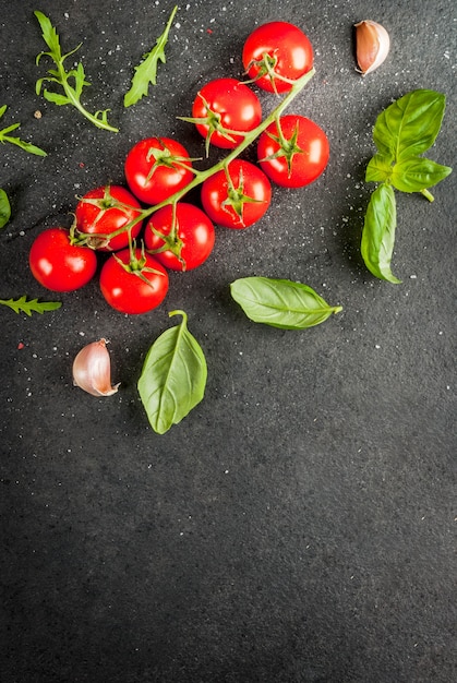 Koken, oppervlak. Ingrediënten voor het koken. Specerijen (zout peper) greens (rucola peterselie basilicum) en cocktail cherry tomaten op een zwarte stenen tafel. Bovenaanzicht kopie ruimte