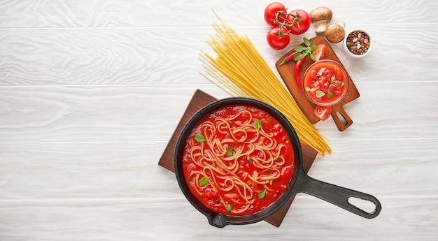 Koken spaghetti met gekookte tomatensaus in gietijzeren pan geserveerd met chilipeper, verse basilicum, kerstomaatjes en kruiden over witte textuur houten tafel, ingrediënten food concept