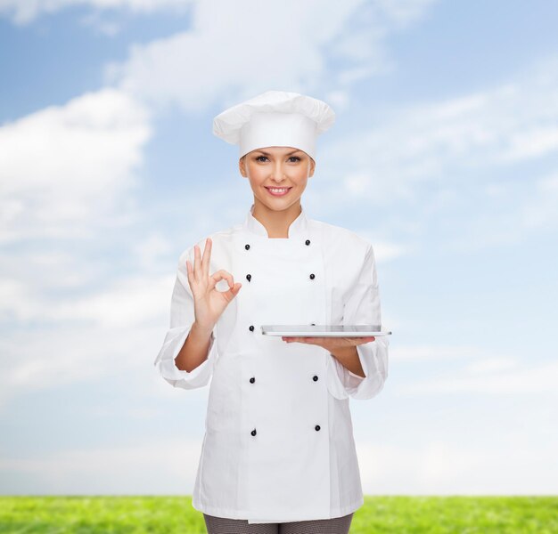 koken, technologie en mensen concept - glimlachende vrouwelijke chef-kok, kok of bakker met tablet pc-computer over blauwe lucht en gras achtergrond