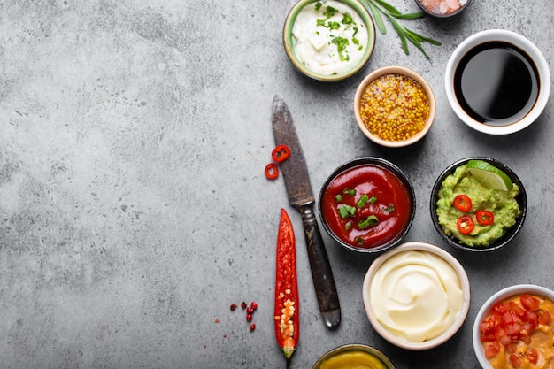 Koken van gezonde sauzen en dressings concept met kopieerruimte. Verschillende dips in kommen op rustieke betonnen ondergrond, ingrediënten, kruiden, peper chili en keukenmes, bovenaanzicht, close-up sjabloon