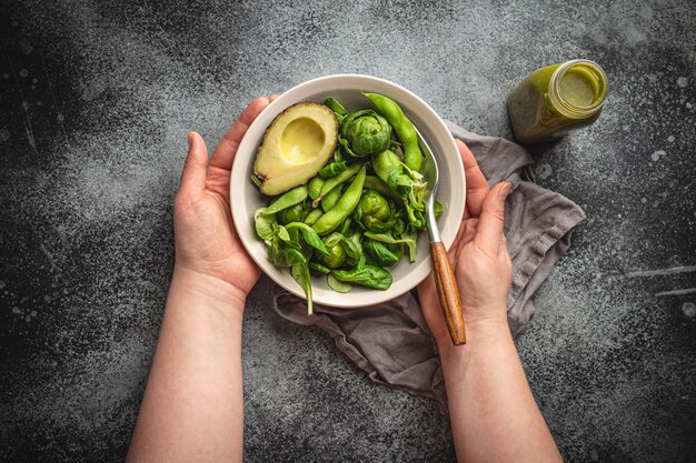 Kom gezonde salade in vrouwelijke handen van bovenaf en groene detox smoothie in een fles op grijze stenen achtergrond, bovenaanzicht. Vrouw eet smakelijke veganistische maaltijd, raw food-dieet en concept voor schoon eten