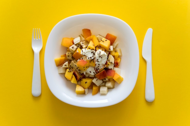 Kom gezonde salade van vers fruit op geel. Gezonde voedingsfruitsalade met zachte kaas en lijnzaad in witte plaat op gele achtergrond met wit mes en vork.