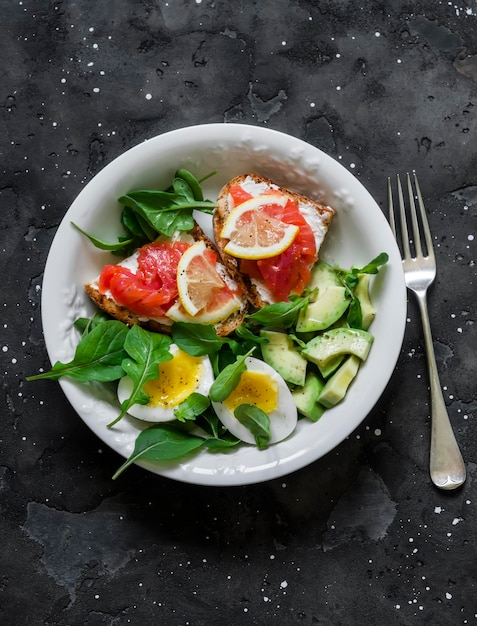 Kom groene salade avocado gekookt ei en sandwiches met roggebrood roomkaas en zalm op een donkere achtergrond bovenaanzicht Heerlijke en gezonde ontbijtbrunch