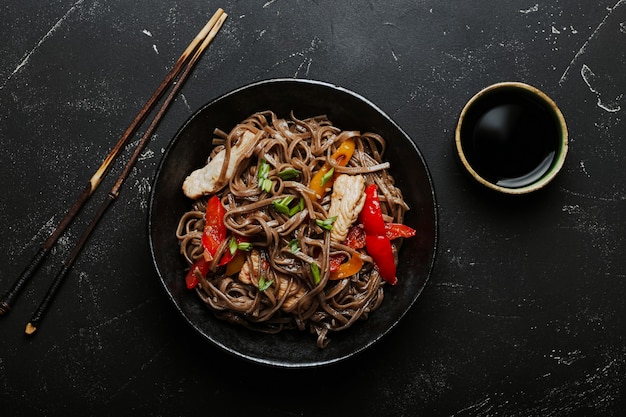 Kom met Aziatische kip roerbak sobanoedels met groenten op donkere zwarte stenen achtergrond van bovenaf, Chinese Thaise of Japanse noedelschotel met sojasaus