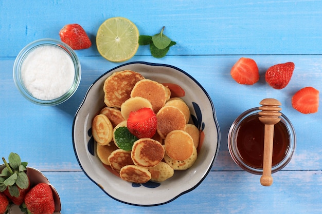 Kom met kleine pannenkoek granen met aardbeien en muntblaadjes op een blauwe houten achtergrond. Trendy eten. Mini cornflakes pannenkoeken.