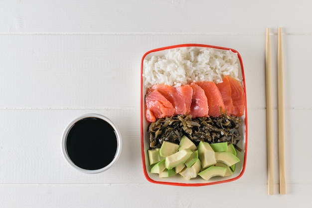 Kom met langkorrelige rijst, zeewier, plakjes avocado, zalm en houten stokken op een lichte tafel. Het uitzicht vanaf de top.