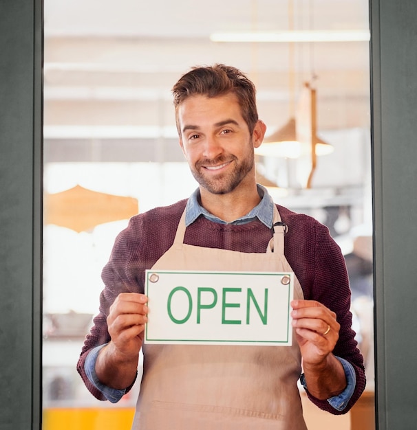 Kom op portret van een jonge ondernemer die in de deuropening van zijn koffiewinkel staat met een open bord