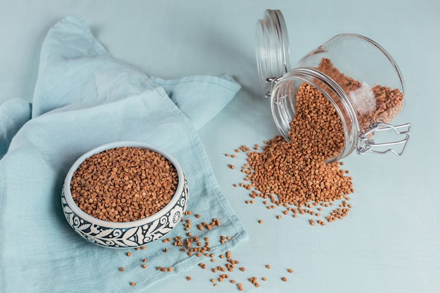 Kom van droge ruwe boekweitgrutten en glazen pot op een lichtblauwe achtergrond
