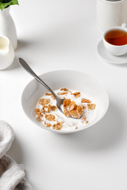 Kom volkoren cornflakes met melk op een witte tafel met een kopje thee. Ontbijt tijd. Witte fotografie.