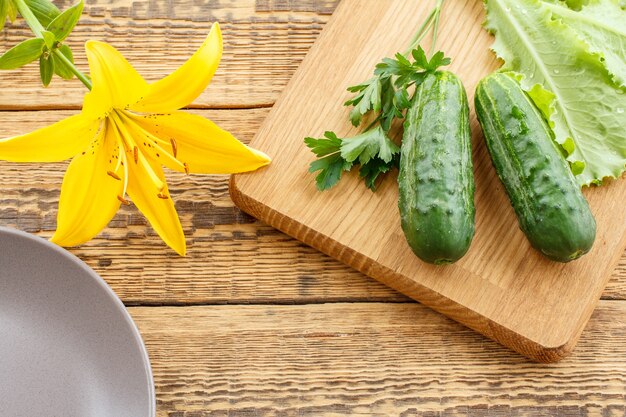 Komkommers en groene salade op snijplank, bord en gele leliebloem op oude houten planken. Net groenten geoogst. Bovenaanzicht.