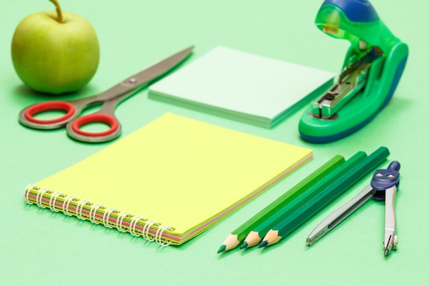 Kompas, kleurpotloden, notitieboekje, notitiepapier, nietmachine, appel en schaar op groene achtergrond.