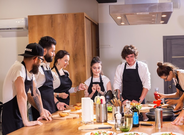 Kookcursus senior mannelijke chef-kok in kookuniform leert jongeren kooklesstudenten om ingrediënten voor gerechten in de keuken van het restaurant te bereiden en te mixen