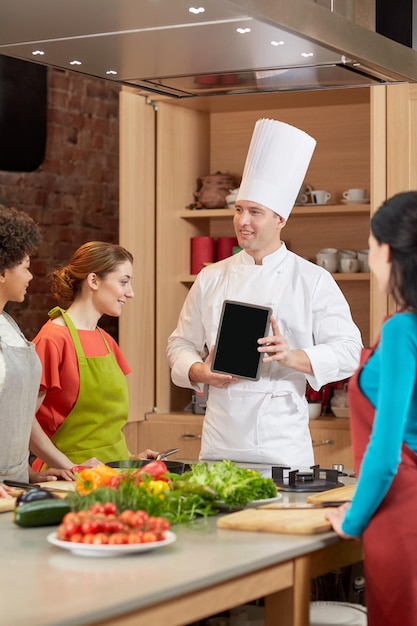 kookles, culinair, eten, technologie en mensenconcept - gelukkige vrouwen met chef-kok die een leeg tablet-pc-scherm in de keuken toont