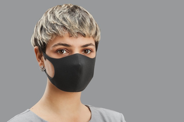 Kop en schouders portret van hedendaagse kortharige vrouw die een masker draagt terwijl ze tegen een minimale grijze achtergrond staat, kopieer ruimte