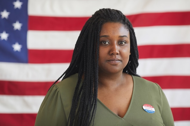 Kop en schouders portret van jonge Afro-Amerikaanse vrouw met STEM sticker terwijl staande tegen de Amerikaanse vlag op verkiezingsdag, kopieer ruimte