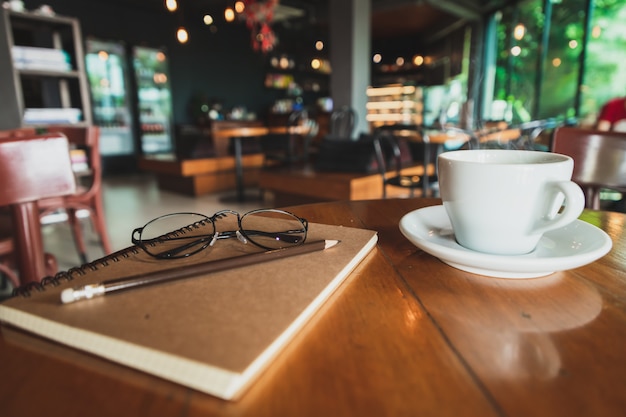 Kop van de close-up de witte koffie, potlood, bruin dekkingsnotitieboekje gezet op een bruine houten lijst in een koffiewinkel