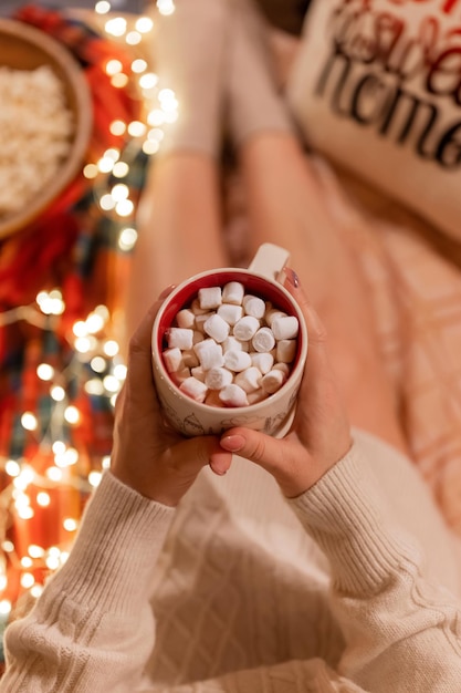 Kopje cacao of koffie drinken met marshmallows in vrouw hand in hand over de benen van vrouw in gebreide trui ontspannen en genieten van de vakantie in gezellig bed thuis op kerst- of oudejaarsavond