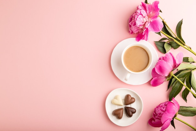 Kopje cioffee met chocolade snoepjes, roze pioen bloemen op roze pastel achtergrond. bovenaanzicht,