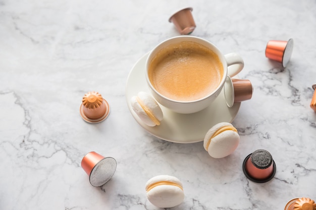 Kopje espresso koffie geserveerd met bitterkoekjes en capsules op marmeren tafel.