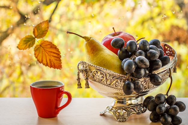 Kopje koffie en metalen vaas met gele peer, tros druiven en appel. Droog blad op vensterglas met waterdruppels en herfstbomen op de achtergrond.