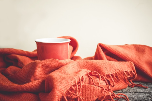 Kopje koffie gewikkeld in een oranje sjaal op een houten tafel en een grijze achtergrond.