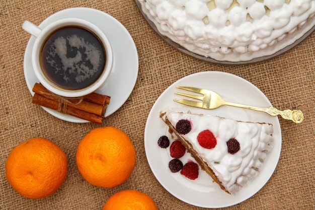 Kopje koffie, kaneel, vork en plakje biscuitcake versierd met slagroom en frambozen, sinaasappels op tafel met zak. Bovenaanzicht.