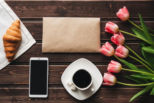 Kopje koffie met een croissant voor het ontbijt op de tafel versierd met een boeket van roze tulpen en een smartphone