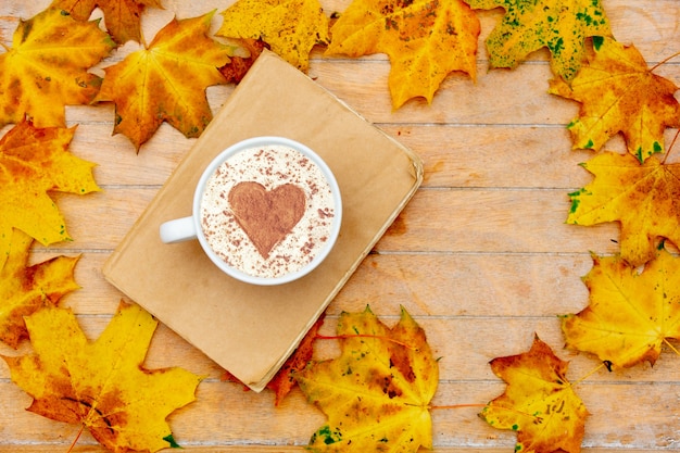 Kopje koffie met een hart van kaneel en boek op tafel, esdoornbladeren rond