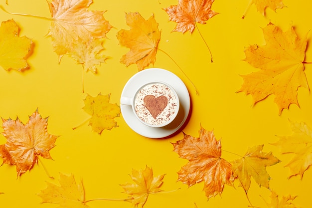 Kopje koffie met hartvorm en esdoornbladeren rond op gele achtergrond.