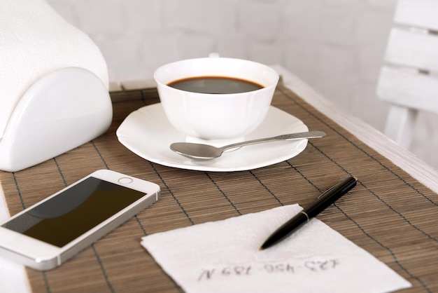 Kopje koffie met mobiele telefoon pen en telefoonnummer op servet op tafel met bamboe mat en witte muur achtergrond