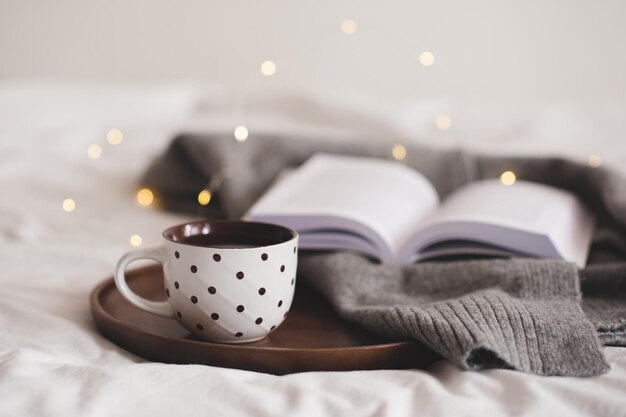 Kopje koffie met stippenpatronen die op een houten dienblad blijven met gebreid textiel en een open papieren boek over gloeiende lichten in de kamer close-up Goedemorgen