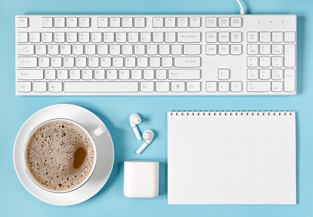 Kopje koffie, toetsenbord, draadloze hoofdtelefoons en Kladblok op lichtblauwe ondergrond.