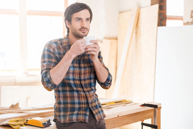 Kopje koffie voor goed werk. Doordachte jonge mannelijke timmerman die een koffiekopje vasthoudt terwijl hij aan de houten tafel leunt met diverse gereedschappen die erop liggen