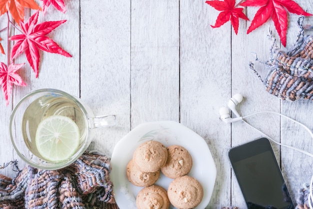 Kopje thee, cookie, sjaal, slimme telefoon en esdoorn bladeren op houten tafel.